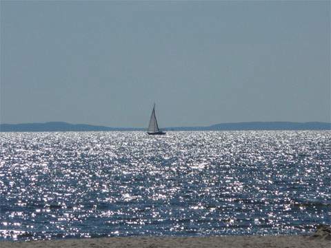 Wie flüssiges Metall: Lichtreflexe auf dem Wasser der Ostsee umgeben das entfernte Segelboot.